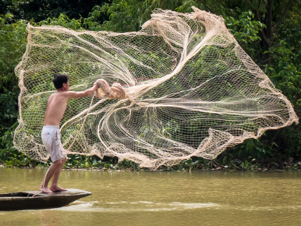 Fisherman casting open net on water from boat in Nihn Bihn 